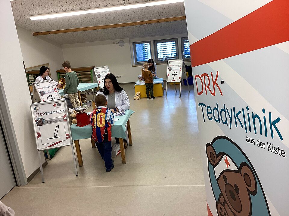 Man blickt in den Raum, in dem die Teddyklinik aufgebaut ist. Am rechten Bildrand steht ein Roll-Up der DRK-Teddyklink. Links im Bild steht ein Tisch. Davor steht ein Flipchart, auf dem "Anmeldung" steht. Ein Junge in einem Fußballtrikot steht vor dem Tisch. Auf der anderen Seite sitzt eine Auszubildende im weißen Kittel. Die beiden sind im Gespräch. Im Hintergrund sind noch andere Stationen der Teddyklinik zu sehen. 