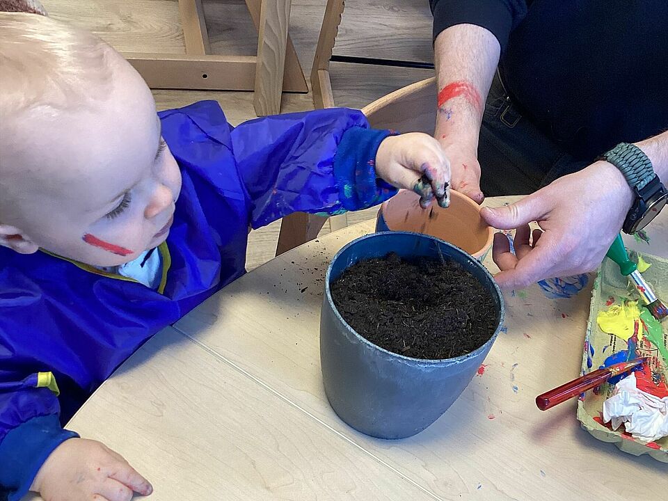 Ein Kleinkind sitzt am Tisch und streut Kressesamen auf die Erde im Tontopf.