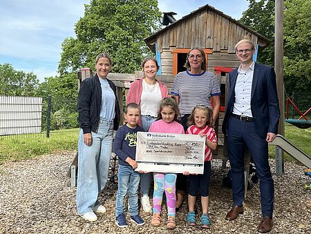 Vier Erwachsene und drei Kinder stehen draußen auf einem Feld. Die drei Kinder im Kitaalter halten einen Scheck in der Hand, die Erwachsenen stehen dahinter. Links eine Dame des Kreisportbundes, ganz recht ein Vertreter der Volksbank, in der Mitte die Kitaleitung und ihre Stellvertreterin. Alle schauen fröhlich in die Kamera.