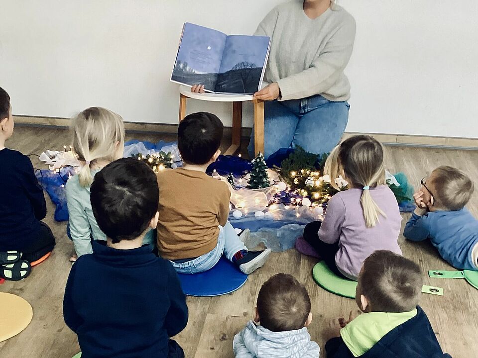 Die Kinder sitzen auf dem Boden und schauen alle in eine Richtung. Vor der Wand sitzt eine Erzieherin, deren Gesicht nicht zu sehen ist. Sie hält das Buch "Die Schneeflocke" in der Hand. Dieses ist aufgeschlagen, so dass alle Kinder die Seiten während des Vorlesens sehen können. Die Kinder sind alle von hinten zu sehen. 