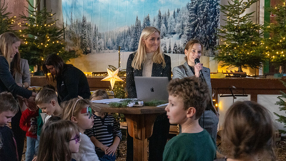 DSC9356 Zwei Frau, die an einem Rednerpult mit Mikrofon stehen und Menschen etwas erzählen. Man sieht einen weihnachtlich geschmücktes Bühnenbild und Kinder in dem Bild