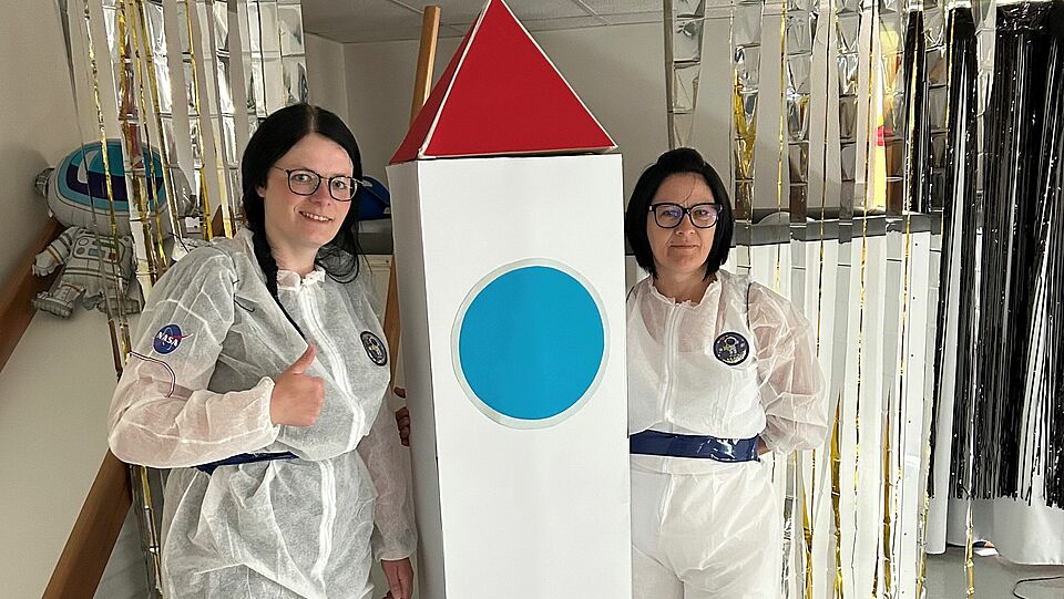 zwei Erzieherinnen als Astronautinnen verkleidet mit einer Rakete zeigen den Daumen hoch