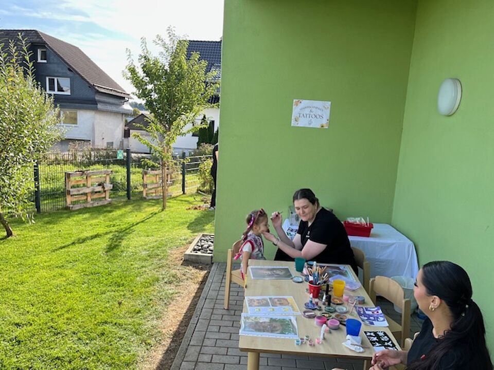 Zwei Erzieherinnen sitzen an einem Tisch, auf dem Schablonen und Farben zum Kinderschminken liegen. Ein Kind wird gerade geschminkt.