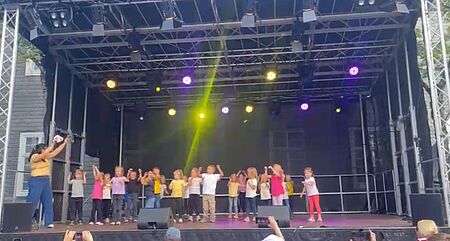 Die Bühne ist in der Totalen fotografiert. Die Gruppe der Kitakinder ist zu sehen, die gerade ihren eingeübten Tanz aufführen. Links neben der Gruppe steht eine erwachsene Frau, die mittanzt und den Kindern Hilfestellung gibt. Im Vordergrund sind Hände mit Handys zu sehen, die aus dem Publikum sind.