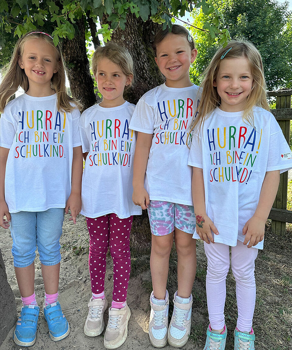 Vier Vorschulkinder posieren nebeneinander mit ihren Abschluss-T-Shirts für ein Foto und lachen fröhlich in die Kamera. 