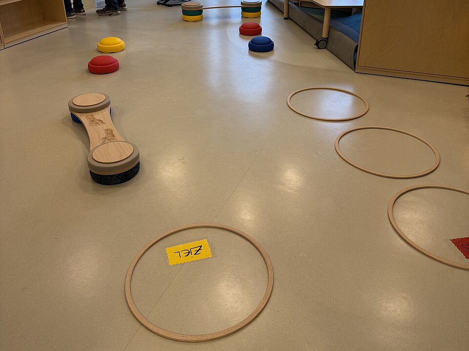 6873 Auf dem Boden sind Ringe und andere Hindernisse zusehen. In einem Ring steht das Wort "Ziel". Es handelt sich um einen Parkour, den die Kinder durchqueren müssen.