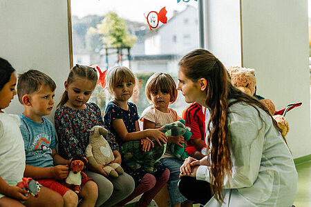 Mehrere Kitakinder sitzen in einer Reihe auf einer Bank, eine Ärztin, die Teddyärztin, kniet vor den Kindern und ist mit ihnen im Gespräch.