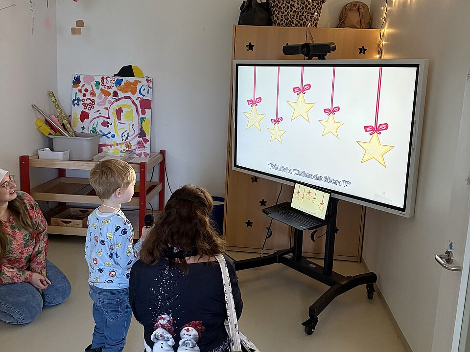 6862 Ein Kind und zwei Frauen hocken vor einem Bildschirm. Das Kind hat ein Mikrofon in der Hand. Auf dem Bildschirm ist ein Liedtext zu sehen.