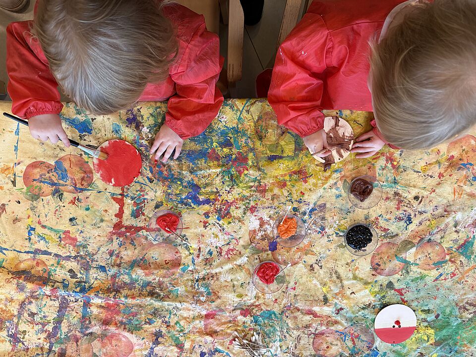 6871 Zwei Kinder bemalen im Kittel Holzscheiben für Weihnachten. Der Tisch ist vor lauter Farben schon ganz bunt.