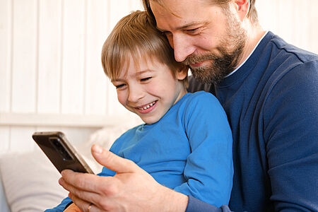 Vater und Sohn schauen gemeinsam fröhlich in ein Handy. Der Sohn sitzt auf dem Schoss des Vaters.