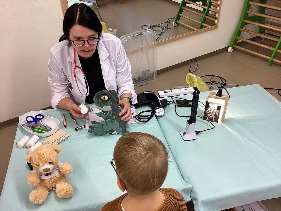 Das Bild ist von oben fotografiert. Ein Junge steht vor einem Tisch, an dem die Teddy-Ärztin sitzt. Sie hat einen weißen Kittel an und Stethoskop um den Hals. In der Hand hält sich einen Kuscheldrachen. Auf dem Tisch liegen Verbände, eine Nierenschale mit einer Schere, ein Blutdruckmessgerät und ein kleines Röntgengerät für Kuscheltiere.