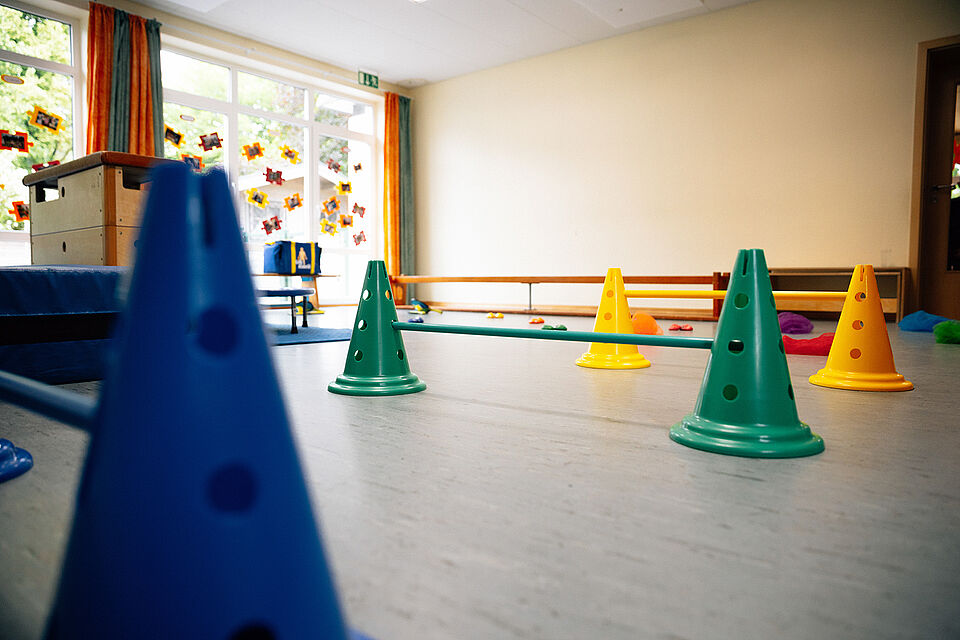 Im Vordergrund steht eine blaue Hürde, dahinter stehen eine grüne und gelbe Hürde. Links ist ein großer Kasten zu sehen. Im Hintergrund steht eine Bank vor einer hellen Wand. Links davon ist eine große Fensterfront.