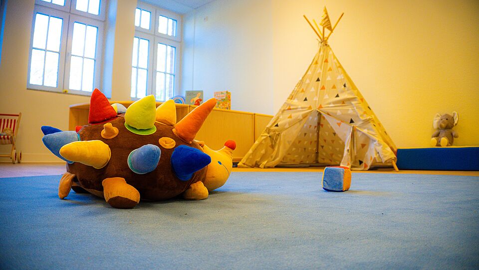 Ein Raum mit blauem Teppich. Auf dem Boden liegt ein großes Stofftier mit bunten, weichen Hörnern. Im Hintergrund steht ein Spielzelt. Daneben sitzt ein grauer Stoffelefant. Es gibt Regale mit Kinderbüchern und große Fenster an der Wand.