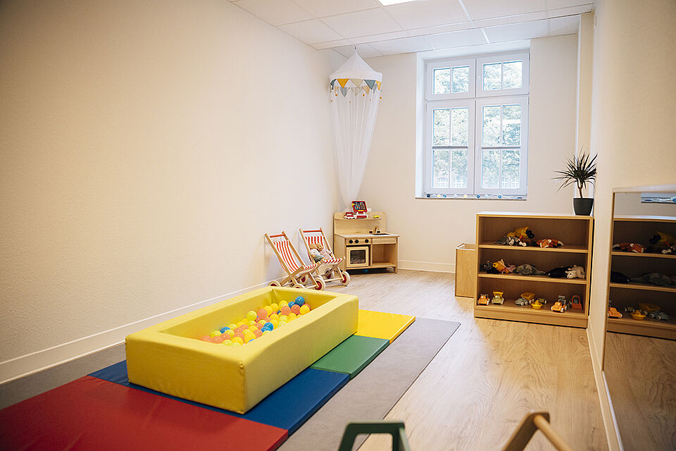 Man schaut in einen Nebenraum für U3-Kinder. An der Wand liegt eine bunte Turnmatte und darauf steht ein kleines rechteckiges Bällebad aus Schaumstoff. Im Hintergrund auf der rechten Seite steht ein kleines Regal mit Spielzeug. In der Ecke des Raums hängt ein kleiner Baldachin.