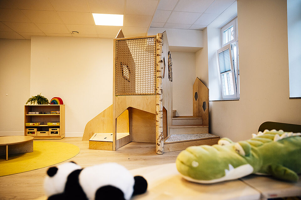 Im Vordergrund liegen eine Plüschpanda und ein großes Plüschkrokodil. Im Hintergrund an der Wand steht ein Klettermöbel aus Holz, daneben ein kleines Regal.