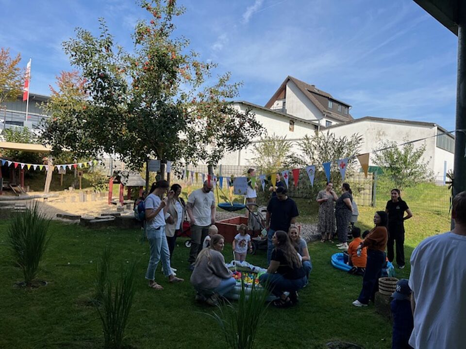 Mehrere Erwachsene stehen und hocken um ein kleines Planschbecken, in dem sich Enten zum Angeln befinden, und unterhalten sich angeregt. Zwei Kleinkinder versuchen, die Enten zu angeln. Weitere Kinder und Erwachsene befinden sich auf der Wiese im Schatten und unterhalten sich.