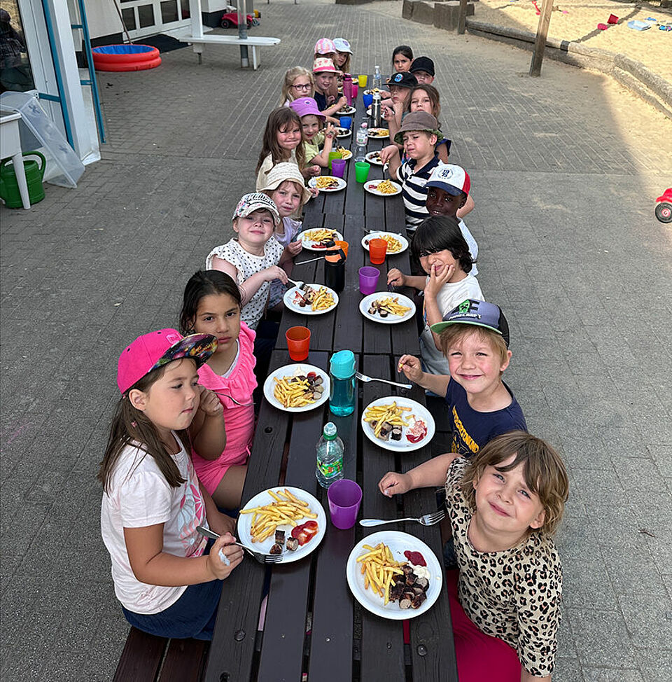 Alle Kinder sitzen draußen bei schönem Wetter am Tisch und essen Pommes und Würstchen.