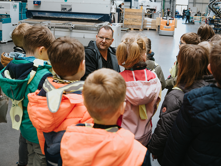Kinder der Kita-Gruppe mit einem Mitarbeiter von Rembe in einer Produktionshalle. Die Kinder stehen mit dem Rücken zur Kamera. Der Mitarbeiter hockt vor den Kindern und sieht sie an.