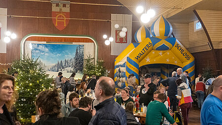 Eine weihnachtlich geschmückte Schützenhalle mit vielen Menschen, die sich in der Halle verteilt haben. In einer Ecke sieht man eine riesige gelbe Hüpfburg