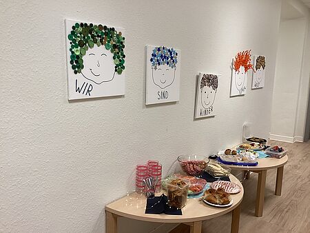 Im Flur der Kita stehen vor einer Wand zwei halbrunde Tische, auf denen ein Frühstücksbuffet aufgebaut ist. Es gibt Brötchen, Gebäck, Rohkostgemüse, Käsespieße und viele andere kleine Leckereien. Über dem Buffet hängen fünf Leinwände. Auf jeder Leinwand ist ein Gesicht gezeichnet, die Haare sind aus Knöpfen, Pfeifenreiniger, Korken, Sternenaufklebern und  kleinen Steinchen. Auf den Bildern steht der Satz: Wir sind Kinder einer Welt. 