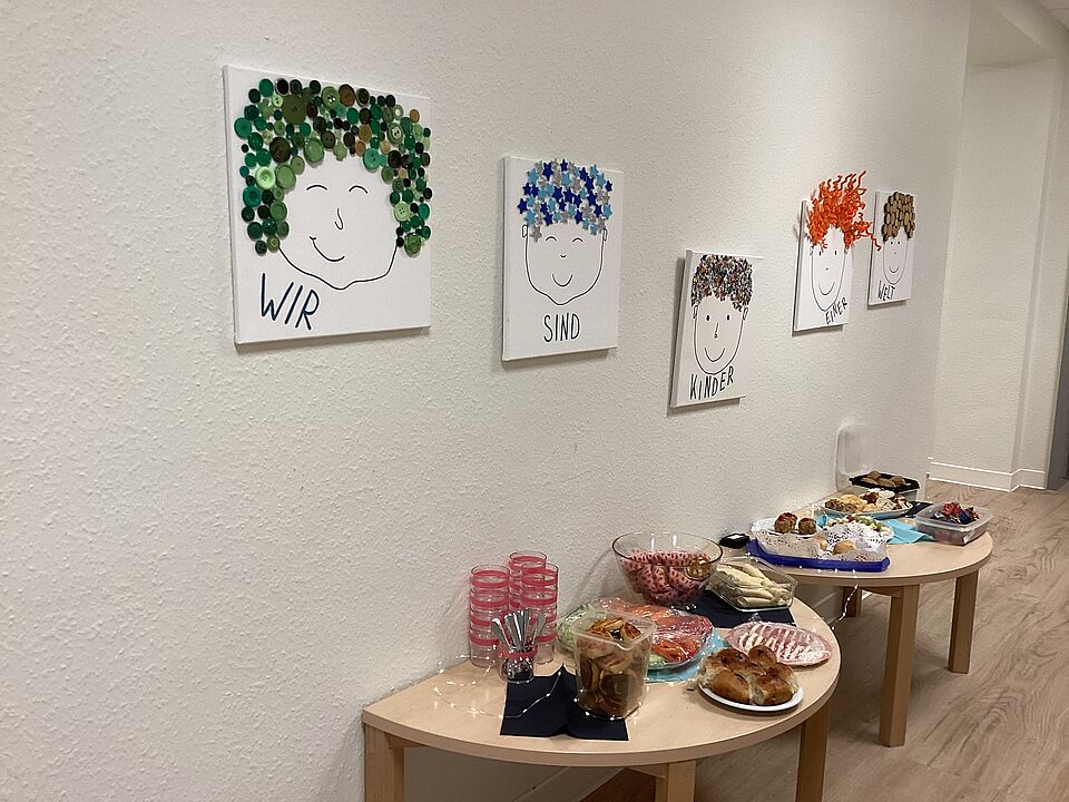 Im Flur der Kita stehen vor einer Wand zwei halbrunde Tische, auf denen ein Frühstücksbuffet aufgebaut ist. Es gibt Brötchen, Gebäck, Rohkostgemüse, Käsespieße und viele andere kleine Leckereien. Über dem Buffet hängen fünf Leinwände. Auf jeder Leinwand ist ein Gesicht gezeichnet, die Haare sind aus Knöpfen, Pfeifenreiniger, Korken, Sternenaufklebern und  kleinen Steinchen. Auf den Bildern steht der Satz: Wir sind Kinder einer Welt. 