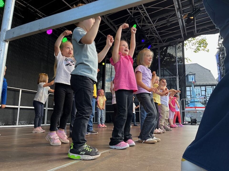 Die Kinder der Kindertageseinrichtung stehen auf der Bühne. Sie sind von unten, von der Seite der Bühne fotografiert. Die Kinder sind alle sportlich gekleidet und heben die Arme.