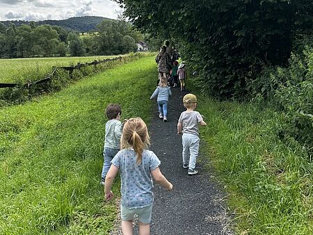 Kinder laufen hintereinander einen Wanderweg entlang. Sie tragen sommerliche Kleidung. Rechts stehen Bäume, links ist ein grünes Feld.