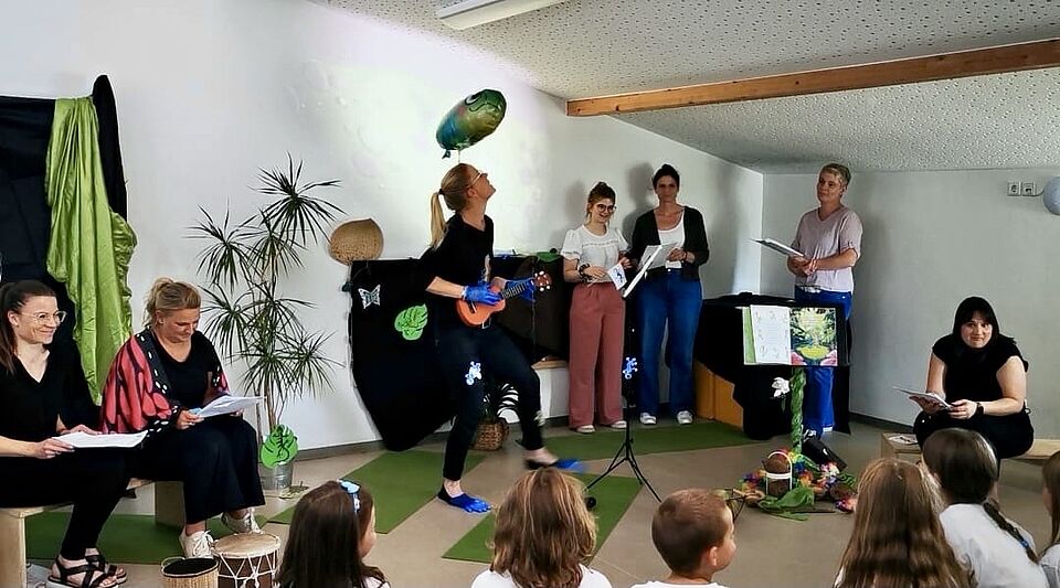 Es stehen mehrere Erzieherinnen auf Fläche, eine hat eine Ukulele in der Hand und köpft einen Luftballon. Die anderen schauen zu. Die Kinder sitzen mit dem Rücken zur Kamera.