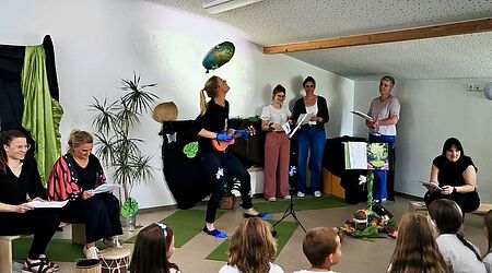 Es stehen mehrere Erzieherinnen auf Fläche, eine hat eine Ukulele in der Hand und köpft einen Luftballon. Die anderen schauen zu. Die Kinder sitzen mit dem Rücken zur Kamera.