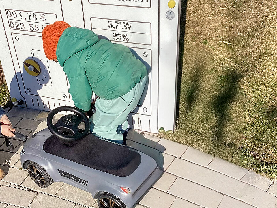 Ein Kind steht an der Tankstelle mit dem Rutschauto und guckt nach dem vorderen Reifen. 