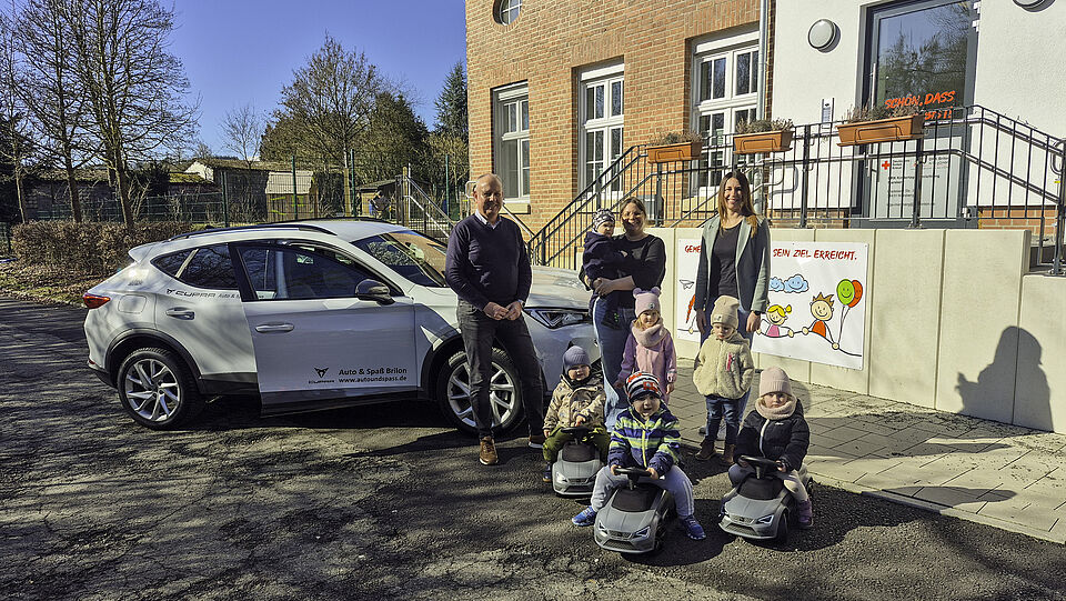 Drei Kinder aus der Kita sitzen auf den neuen Rutschautos von CUPRA, dahinter stehen zwei weitere Kinder und drei erwachsene Personen. Rechts stehen zwei Frauen, links vor einem weißen CUPRA ein Mann, der etwas älter ist als die beiden Frauen. Alle gucken direkt in die Kamera. Die Sonne scheint uns im Hintergrund ist das Gebäude der Kita mit Eingang zu sehen.