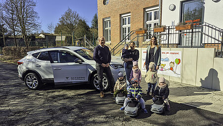 Drei Kinder aus der Kita sitzen auf den neuen Rutschautos von CUPRA, dahinter stehen zwei weitere Kinder und drei erwachsene Personen. Rechts stehen zwei Frauen, links vor einem weißen CUPRA ein Mann, der etwas älter ist als die beiden Frauen. Alle gucken direkt in die Kamera. Die Sonne scheint uns im Hintergrund ist das Gebäude der Kita mit Eingang zu sehen.