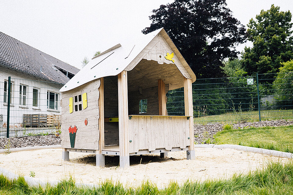 Im Sand steht ein großes Spielhaus aus Holz. Es ist vorne halb offen und ist mit Blumen bemalt.